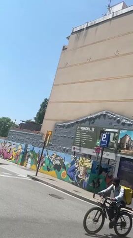 The video shows a street in a town. The camera appears to be moving, possibly from a car or bicycle. Initially, a large building with a mural depicting characters from Dragon Ball can be seen. A parking sign and a Taco Bell advertisement are also visible. The camera then pans to the right, showing a building with a motorcycle parked in front.  Further down the street, there are people walking and sitting on benches. The street has trees lining both sides, forming a green canopy overhead. A red and white zebra crossing is visible on the road with a speed limit sign of 20. Several cars are parked along the street, and a few are driving. The day appears to be sunny.