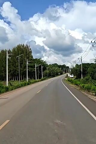 This video shows a perspective from a moving vehicle on a paved road. The road has a yellow line marking the center and a white line marking the right edge. On the left side of the road, there are trees and tall poles with lights. On the right side of the road, there are more trees, a few buildings with reddish roofs, and utility poles with wires. The sky is blue with large, white, fluffy clouds. The video captures the scenery as the vehicle continues down the road.