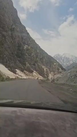 The video shows a car driving on a paved road through a mountain pass. A large rocky mountain rises on the left side of the road, with snow patches at its base. In the distance, snow-capped mountains are visible. A few other vehicles can be seen further down the road. The sky is blue with scattered clouds.  Music is playing in the background. The video is shot from the driver's perspective, looking out the windshield.
