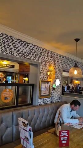 The video shows the interior of a pub or restaurant. Several people are seated at tables, looking at menus or using their phones. A man in a white jacket is looking at his phone. Another man wearing a yellow high-visibility jacket is looking at a menu. A few other tables are occupied with groups of people. There is a TV playing in the background. The decor includes patterned wallpaper, pendant lights, and framed pictures on the walls. The room has a casual and relaxed atmosphere.