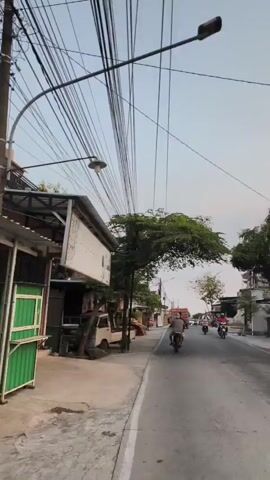 The video shows a street scene with a clear sky. On the left side of the road, there is a building with a sign that reads "Griya Batik". In front of the building, there are stacks of what appear to be wooden planks and a white truck. Several trees line the sidewalk. The road itself has a few motorcycles and a truck driving on it. The street is lined with more buildings and trees, with numerous overhead power lines stretching across the frame. The overall impression is of a commercial area in a town or city.