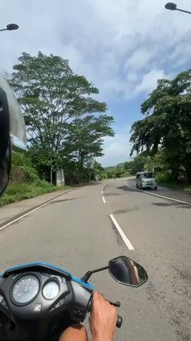 A first-person perspective video shows someone riding a blue and black scooter on a paved road. The rider wears a helmet, visible on the left side of the frame. The road has a solid white line down the center. Lush green trees line both sides of the road. The sky is partly cloudy with patches of blue. The scooter's speedometer and handlebars are visible in the foreground. A rearview mirror shows the rider's reflection. In the distance, another scooter can be seen on the road. Streetlights are sporadically placed along the roadside. The overall environment appears to be a tropical or subtropical setting. The video has a somewhat shaky quality, likely due to the movement of the scooter.