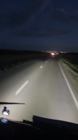 The video shows a first-person perspective of driving on a highway at night. The headlights of the vehicle illuminate the asphalt road ahead, with white lines marking the lanes. The sky is dark blue with scattered clouds, and in the distance, city lights and the headlights of other vehicles are visible, suggesting the approach to a populated area. The scene is set in Baiqadam, Kazakhstan. There are no people clearly visible, but the presence of a dashcam on the dashboard indicates the viewpoint is from inside a vehicle, likely a car or truck. The overall mood is one of travel and transition, with the darkness of night contrasting with the distant lights and the illumination provided by the vehicle's own headlights. The weather appears clear, with no rain or fog. No text is visible in the video.