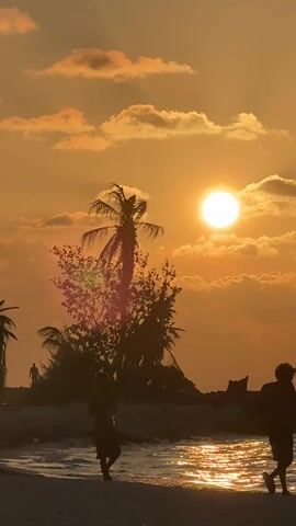The video shows a silhouetted figure, possibly a child, standing on a beach at sunset. The sun is prominent in the sky, partially obscured by scattered clouds, casting an orange glow over the scene. The ocean reflects the sunlight, creating a shimmering effect on the water's surface. Palm trees and other vegetation are silhouetted against the bright sky. A few other figures are visible in the distance on the beach. The camera is stable, capturing the static scene.