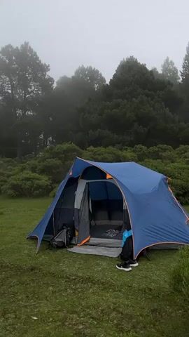 A blue tent is pitched on a grassy clearing surrounded by trees. The sky is overcast and foggy, and the overall atmosphere is damp and cool. There is camping equipment near the tent, including chairs and a table. In the background, more trees and vegetation are visible. The scene suggests a remote camping location.