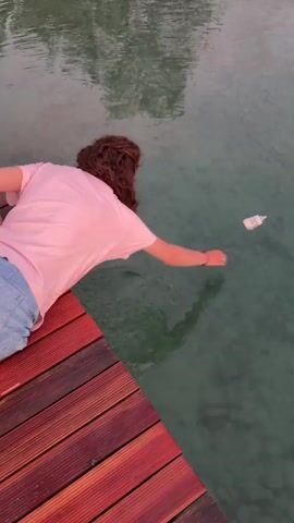 A person with long, curly brown hair, wearing a pink t-shirt and denim shorts, is lying on a wooden dock. Their body is partially over the edge of the dock, with their arms reaching towards the water. A white baby bottle is floating in the clear, greenish water a short distance away from the person. The person appears to be trying to reach or retrieve the bottle. The sun is low in the sky, casting a warm glow on the scene and creating a soft, pinkish hue in the water. The overall atmosphere suggests a peaceful, late afternoon or early evening setting. The scene is set at Lake Hallwil in Meisterschwanden, Switzerland. No visible text is present in the media.