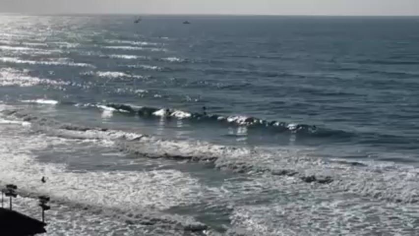 The video shows a wide shot of a coastline on a bright, sunny day. The ocean stretches into the horizon, with several small boats visible in the distance. Closer to the shore, waves are breaking, creating white foam as they roll in. A few people are visible in the water, some appearing to be surfers riding the waves. In the lower left corner, a portion of a building with lights attached can be seen.