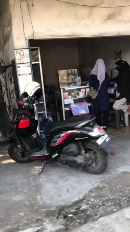 The video shows a street scene with a red and black scooter parked on the left, facing right. A white helmet is on the handlebars. Behind the scooter is a storefront with a partially open door and a window. Inside the store, two women wearing hijabs are visible. One woman in a blue hijab is standing at a counter, and another woman in a black hijab is also at the counter. There are shelves and displays of products inside the store. A white stool is near the counter. The ground in front of the store is concrete and appears somewhat worn. The building's exterior wall is visible and seems to be painted a light color.