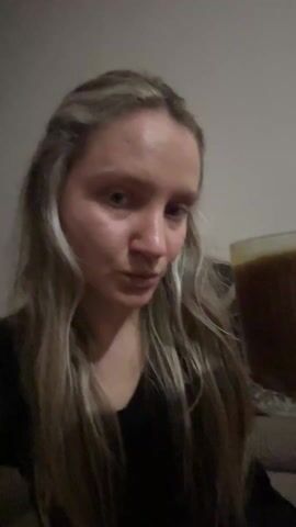 Here's a detailed description of the video:

A young woman with long blonde hair is holding a large glass mug filled with a dark brown liquid. She’s wearing a black top and has a nose ring. She brings the mug to her lips, takes a sip, and makes a "Mmm" sound, indicating she enjoys the drink. She then smiles slightly and raises the mug in a toasting gesture towards the camera. The background appears to be a plain wall, possibly in a home setting.