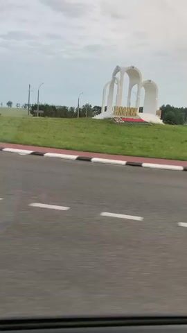 A white arch monument with the Cyrillic word "НАСТАСЬ" in gold letters stands on a grassy hill. The monument has three archways of varying heights. In the foreground, a red and white striped barrier is visible, along with a road with white lines. The sky is overcast. The video appears to be taken from a moving vehicle.