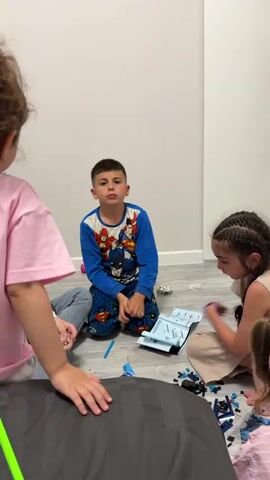 A young boy in pajamas, patterned with superhero designs, sits on a light grey wooden floor, and is seen doing a pouting face directly to the camera. Two other people are partially visible in the frame on either side of him. A blue marker rests on the floor near the boy. A light blue folded paper object is visible near the lower right person. The boy sits in front of a plain white wall.