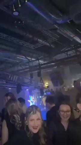 The video shows a crowd of people in a dimly lit, industrial-looking building with exposed pipes and lighting rigs on the ceiling. Many people are dressed in dark clothing.  There are blue and yellow stage lights in the background, possibly illuminating a stage or performance area. The camera is held by a person who appears briefly at the edge of the frame. Several women are visible in the foreground, some laughing and interacting with each other. One woman briefly raises her middle finger towards the camera.  The overall atmosphere suggests a club or event with music and social interaction.