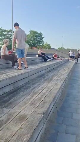 The scene shows a group of people near a beachside area on a sunny day. The camera is positioned at a low angle, looking across a set of wooden steps towards a paved walkway, with a sandy beach visible in the background. Several people are sitting and lounging on the wooden steps, enjoying the weather. A group of people, mostly men, are standing together on the paved walkway, some facing the camera, others talking among themselves. In the background, more people are visible on the beach, along with structures like a tent or canopy. The sky is clear and blue, indicating a pleasant day. The walkway is made of grey stone bricks.
