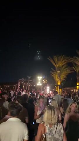 A crowded outdoor venue at night. People are gathered closely together, facing towards the center of the space. Numerous lights are visible, including string lights, spotlights, and larger, multi-bulb fixtures mounted overhead. A palm tree with illuminated fronds is on the right side of the frame. Some individuals are looking at their phones. The atmosphere appears to be festive and social.