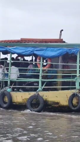 A passenger ferry travels through murky water under an overcast sky. The ferry is a flat-bottomed vessel with a yellow hull and a green railing. It is covered by a blue tarp supported by poles. Several people are visible on the deck, some standing and some seated. Life preservers are hanging on the railing. Tires are attached to the sides of the hull as fenders. A bird is seen flying above the ferry. The water is choppy, indicating the ferry is in motion.