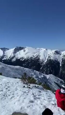 A woman in a red jacket sits on a snowy mountain peak overlooking a range of snow-capped mountains under a clear blue sky. She stands, adjusts what appear to be ropes and gets ready to do something adventurous. An object presumed to be a helmet lays in the snow nearby.