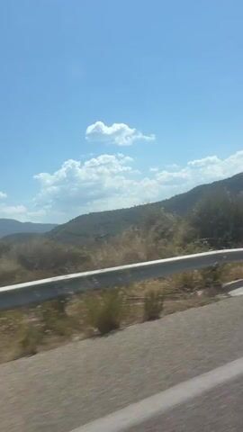 The video shows a view from a moving vehicle, likely a car, on a sunny day. The camera is positioned to capture the scenery passing by on the right side. In the foreground, a gray metal guardrail runs horizontally across the frame, separating the road from the adjacent landscape. Beyond the guardrail, dry, yellowish grass and some sparse bushes are visible. In the background, rolling hills covered with vegetation stretch towards the horizon. The sky is a clear, bright blue with a few wispy white clouds. The scene is likely a rural or mountainous area, possibly a roadside with a view of natural landscapes. The motion blur indicates that the vehicle is traveling at a moderate speed.