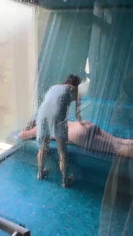 The video shows a person in light clothing standing over a person lying on a blue surface, viewed through a wet, transparent barrier. The person standing appears to be adjusting something near the person lying down. The environment is slightly obscured due to water droplets on the barrier.
