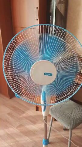 A white and blue oscillating fan is displayed in a room. The fan is in focus in the foreground, with a wooden cabinet and part of a wall visible in the background. A stool is positioned to the right of the fan. The fan is on and its blades are rotating.