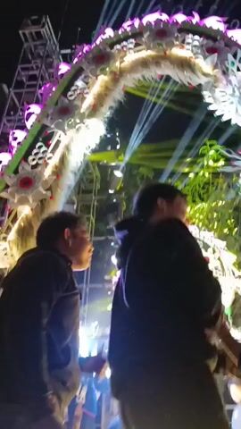 This is a close-up, low-angle video with a shallow depth of field, focusing on the upper bodies of two individuals. They are standing side-by-side, facing away from the camera and towards a brightly lit, decorated archway. The archway is adorned with white and pink floral elements and appears to be part of an outdoor event, possibly a festival or a celebration, given the presence of stage lighting and what looks like a crowd in the background. The two individuals are wearing dark hoodies. The person on the left is looking upwards, while the person on the right is looking towards the right, with their arm extended as if to interact with something or someone off-camera. The lighting is dramatic, with strong beams of light cutting through a hazy atmosphere, suggesting the presence of smoke or fog machines. The overall impression is one of a lively, possibly nighttime event.