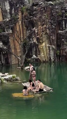 A group of five people are enjoying a day in a quarry with a clear green body of water. Two individuals are in the water, one standing and one partially submerged near a small floating platform. A bamboo raft is in the center of the frame with three individuals seated on it, one of whom is holding a wooden oar. The backdrop is a sheer, rocky cliff face with vertical striations and sparse vegetation.