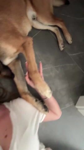 A woman is lying on her back on a gray tiled floor.  She is wearing a white v-neck t-shirt, a gold chain necklace, and has a small earring in her left ear. Her brown dog is lying next to her, with its paws on her arm. The woman looks at the camera and smiles. The dog rests its head on the woman’s head.