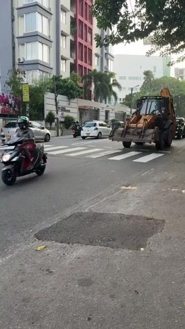 A yellow backhoe loader is parked on a street, its front loader raised. The vehicle is positioned across a pedestrian crossing, blocking traffic. To the left of the backhoe, a white van and a silver car are stopped at the crossing. A motorcyclist is visible between the van and the backhoe, also stopped. In the background, a modern multi-story building with a red facade dominates the scene, with trees and other buildings visible further back. The street is paved, and a pothole is present in the foreground. The lighting suggests it is daytime, possibly late afternoon or early morning.
