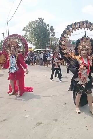 Two women in elaborate costumes are participating in a parade. The woman on the left wears a long, flowing red dress and a large, circular headpiece adorned with red flowers and gold trim. The woman on the right wears a black outfit with a short skirt and a similarly large, circular headpiece decorated with black feathers and gold trim. Both women have ornate sashes draped across their chests, which appear to be made of layered paper or fabric, possibly decorated with images or text. They are walking on a paved street lined with spectators. In the background, there are trees, buildings, motorcycles, and more people watching the procession. The atmosphere appears festive and celebratory.