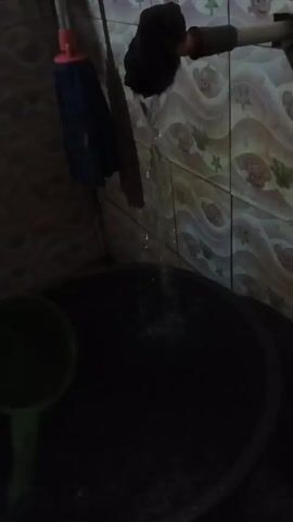 A dark video shows a faucet dripping water into a large dark tub. A green ladle sits in the tub, and a mop with a blue head and pink handle hangs on the wall behind the tub. The wall behind the faucet is tiled with a wavy pattern and images of cartoon characters.