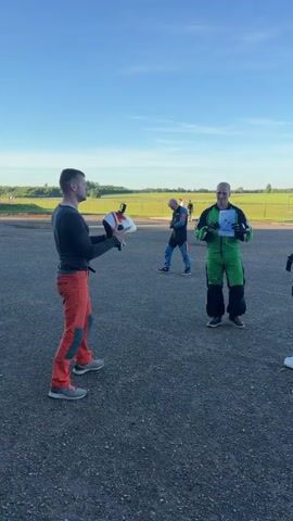 The video opens with a man on the left wearing a black long-sleeved shirt and orange pants holding a white and red object. He is looking to his right, towards a man in a bright green jumpsuit who is holding a certificate or document and smiling. To the right of the man in green is a woman in a blue jumpsuit, looking towards the man in green. In the background, several other people are visible, some in athletic wear, and a field with green grass and a line of trees. The ground is paved. The sky is a clear blue with a few wispy clouds. The camera appears to be handheld and is panning slightly to the right. There is sound of speech throughout the clip.