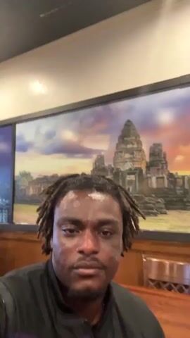 The video shows a dark-skinned man with dreadlocks filming himself in what appears to be a restaurant. He is wearing a black shirt with a purple stripe across the chest. The restaurant has a neutral-colored ceiling with recessed lighting and oriental-style hanging lamps. Other patrons are seated at tables in the background. A large picture hangs on the wall behind the man, depicting an image resembling Angkor Wat at sunset. The camera moves slightly, showing more of the restaurant's interior.