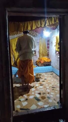 This video shows a man standing in a doorway, looking into a small, dimly lit room. The man is wearing a light-colored shirt and an orange dhoti. He appears to be facing away from the camera and towards an altar or shrine within the room.

The room is decorated with yellow drapes and has a tiled floor. On the altar, there is a small blue platform with a golden cloth draped over it, suggesting it holds a sacred object or deity. A small oil lamp is lit on the floor in front of the altar, casting a warm glow. The walls of the room are white and appear to be made of rough plaster. The overall atmosphere is one of reverence and quiet prayer.

The video seems to capture a moment of religious devotion, possibly during a puja or prayer ritual. The time of day is unclear due to the indoor setting and artificial lighting, but the dim light suggests it could be evening or early morning. There is no visible text in the media file. The location context of Naggar, India, implies that this scene likely takes place in a Hindu temple or a domestic shrine.