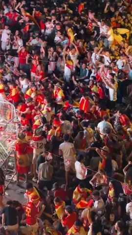 This is a vertical, medium shot video of a large, dense crowd of people outdoors at night. The crowd is predominantly young adults and is very energetic, with many individuals raising their arms in the air and holding small flags. The flags appear to be red and yellow, suggesting a national or team affiliation. The lighting is dim, with some artificial lights illuminating the scene, creating shadows and highlighting the movement of the crowd. There are metal barricades visible in the foreground, indicating a controlled or designated viewing area. The overall atmosphere is one of celebration and excitement.