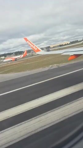 The video shows an airplane on the runway through the window of another plane. The wing of an EasyJet airplane is visible with the company logo in orange. The airplane moves very fast down the runway under a cloudy sky.
