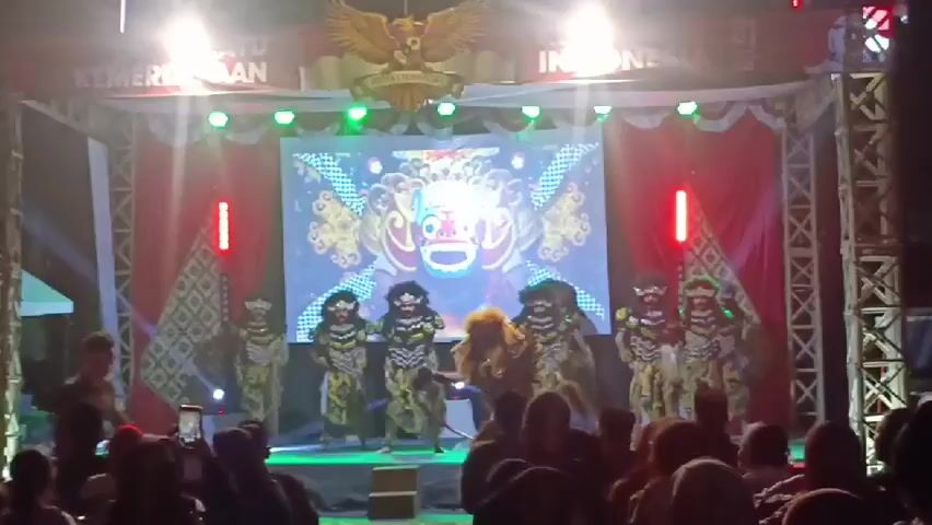 A group of performers in traditional costumes are on a stage, dancing. The performers are wearing masks and elaborate outfits. Behind them, a large screen displays an animated image of a mask. Red and white banners are hung above the stage, with an eagle emblem in the center. Lighting is bright, with spotlights illuminating the stage. An audience is visible in the foreground, with some people holding up their phones to record the performance. The overall setting appears to be an outdoor event or festival.
