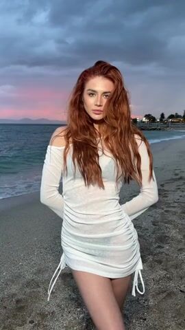 A woman with long red hair stands on a beach, looking towards the camera and posing. She wears a semi-transparent white, off-the-shoulder dress with long sleeves. The beach is made of small pebbles and the sea is behind her. The sky is overcast with hints of pink. She moves her hair with her hand. The scene is set at dusk.
