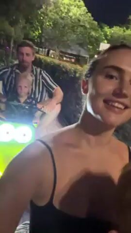 At night, a woman records a video of a man riding a lime green scooter with neon lights on a sidewalk. The man holds a toddler in front of him. The sidewalk is tiled with a light grey repeating pattern. Trees and bushes are visible in the background, along with streetlights. The man wears a striped shirt, and the woman wears a spaghetti strap tank top. She holds the camera toward her, smiling and giggling. The video is blurry.
