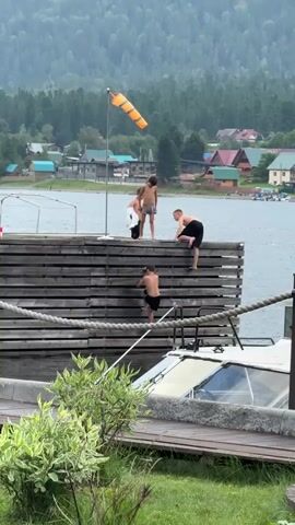 Four boys are on a wooden dock. Two boys are standing on the dock, one boy is climbing over the edge of the dock, and one boy is climbing up the side of the dock. The dock is next to a body of water. In the background, there are trees, houses, and a mountain. There is a windsock on a pole on the dock.