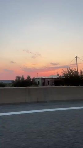 A time-lapse video captures a sunset over a highway. The sky transitions from a light blue to shades of pink, orange, and red as the sun dips below the horizon. Silhouetted trees and distant mountains are visible against the colorful sky. A concrete barrier runs along the side of the highway in the foreground. The video is shot from a moving vehicle, giving a sense of travel.