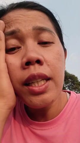 The video shows a close-up of a woman's face as she speaks. Her left hand is supporting her head. She is wearing a pink t-shirt. The background is blurred and appears to be outdoors with a tree visible. The woman's expression seems to convey sadness or distress.