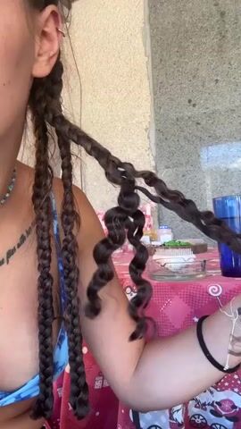 A young woman with braided hair is shown from the chest up, outdoors. She is wearing a blue bikini top. Her hair is dark brown and styled in long braids that are twisted at the ends. She is holding one of her braids with her right hand and is manipulating it, showing off the spiral curls. Her left hand is touching another braid. In the background, there is a table covered with a pink tablecloth featuring a Christmas gnome and train pattern. A blue glass of liquid and some other objects are on the table. The wall behind her is light-colored and textured.