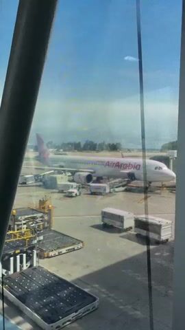 A person is looking out an airport window, showing an Air Arabia plane parked on the tarmac, with other planes visible in the distance. Ground crew and equipment are around the plane. The camera moves left and right, showing more of the airport.