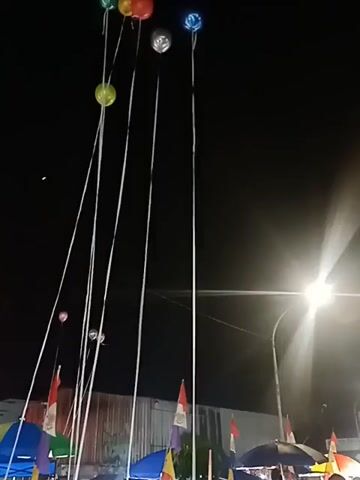 The video captures a night scene with several balloons tied to long strings. The balloons are of various colors, including green, silver, and blue, and are floating high in the dark sky. Below the balloons, there are several colorful umbrellas and flags. The flags appear to be red and white, possibly national flags. There are also some indistinct figures of people visible beneath the umbrellas and tents, suggesting an outdoor event or market. A bright light, possibly a streetlamp, illuminates the right side of the scene, casting rays into the darkness. The overall impression is of a festive atmosphere taking place after dark.