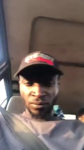 A man with dark skin, a short beard, and a baseball cap is sitting in the front seat of a vehicle, wearing a seatbelt. He is looking directly at the camera. Two other people are visible behind him, one slightly out of focus. The interior of the vehicle appears to be older. The camera is shaky.
