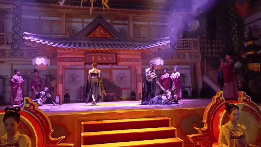 A stage performance is taking place, featuring several actors in traditional Chinese costumes. The stage is set to resemble an ancient Chinese palace or courtyard, complete with wooden architecture, ornate carvings, and lanterns. In the center of the stage, a woman in a black and gold outfit stands prominently, while other actors are positioned around her. Some actors are kneeling or in dynamic poses, suggesting a dramatic or action-filled scene. Two women in yellow and gold costumes are seated on either side of the foreground, framing the stage. The lighting is theatrical, with purple and pink hues illuminating the stage, and smoke effects add to the atmosphere. The overall impression is that of a historical or mythological drama being performed.