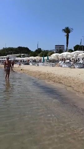 The video shows a sunny day at a crowded beach in Trani, Italy. The scene is set along the shoreline, with the calm, shallow water lapping at the sand. The beach is lined with numerous white umbrellas, providing shade for beachgoers. Loungers and chairs are arranged neatly under the umbrellas, indicating a well-maintained beach establishment.

People are engaged in various beach activities. Some are walking along the water's edge, while others are relaxing on the sand under umbrellas. Children are playing, and a few individuals appear to be sunbathing. A prominent object in the scene is a red lifeguard boat with orange life preservers, positioned near a white lifeguard tower. A lifeguard is visible in the tower, overseeing the beach. A palm tree stands tall against the clear blue sky, adding a tropical feel to the setting. The overall atmosphere appears to be one of leisure and enjoyment, typical of a summer day at the beach.

Notable details include the bright sunlight and clear blue sky, suggesting fair weather. The presence of beach umbrellas, loungers, and a lifeguard station confirms this is a public or private beach area designed for recreation. The text "SALVATAGGIO" is visible on the lifeguard tower, which translates to "rescue" or "lifeguard" in Italian, reinforcing the context. The time of day appears to be midday, given the strong sunlight and the busyness of the beach.