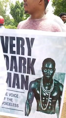 Here is a concise description of the video:

The video shows a man holding up a poster that reads "VERY DARK MAN" in large black letters. Underneath, it says "THE VOICE OF THE VOICELESS". The poster also has a depiction of a dark-skinned man with prominent muscles, wearing necklaces, and a white loincloth. The man holding the poster is wearing a light pink sweater, red slides, and a red baseball cap. He appears to be outdoors, possibly at a rally or gathering, as there are other people and a motorcycle visible in the background.