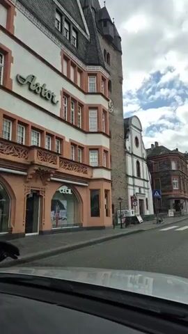 The video shows a first-person perspective from inside a moving vehicle, driving through a European town on a cloudy day. On the left side of the road, there are several multi-story buildings with traditional architecture, some with shops on the ground floor. A large stone tower with a clock face is prominent on the left. Further down the road, there is a white building with a distinctive facade. On the right, a hillside covered with trees and what appears to be a castle or historic structure on top is visible in the distance. The road has painted lane markings and some temporary traffic barriers with red and white stripes. There are also street signs, including a blue circular sign with a white arrow pointing left and a red octagonal STOP sign. Other vehicles and a few pedestrians can be seen along the street. The dashboard of the vehicle is visible at the bottom of the frame, with a silver object (possibly a knife) resting on it.