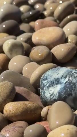 The video shows a close-up of rounded, wet pebbles on a beach. Water splashes briefly over the rocks. The pebbles are in various shades of brown and tan, with one larger rock that is a mottled gray and white. Sunlight glints off the wet surfaces.