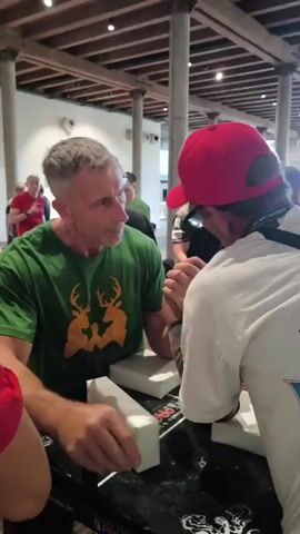 Two men are arm wrestling at a table in what appears to be an indoor event space. The man on the left is wearing a green t-shirt with a logo featuring two deer heads. He has gray hair and is looking intently at his opponent. The man on the right is wearing a white t-shirt and a red baseball cap. Their hands are clasped on the table, and they are both leaning forward, exerting force. Several other people are visible in the background, some watching the arm wrestling match. The environment has wooden ceiling beams and large pillars.